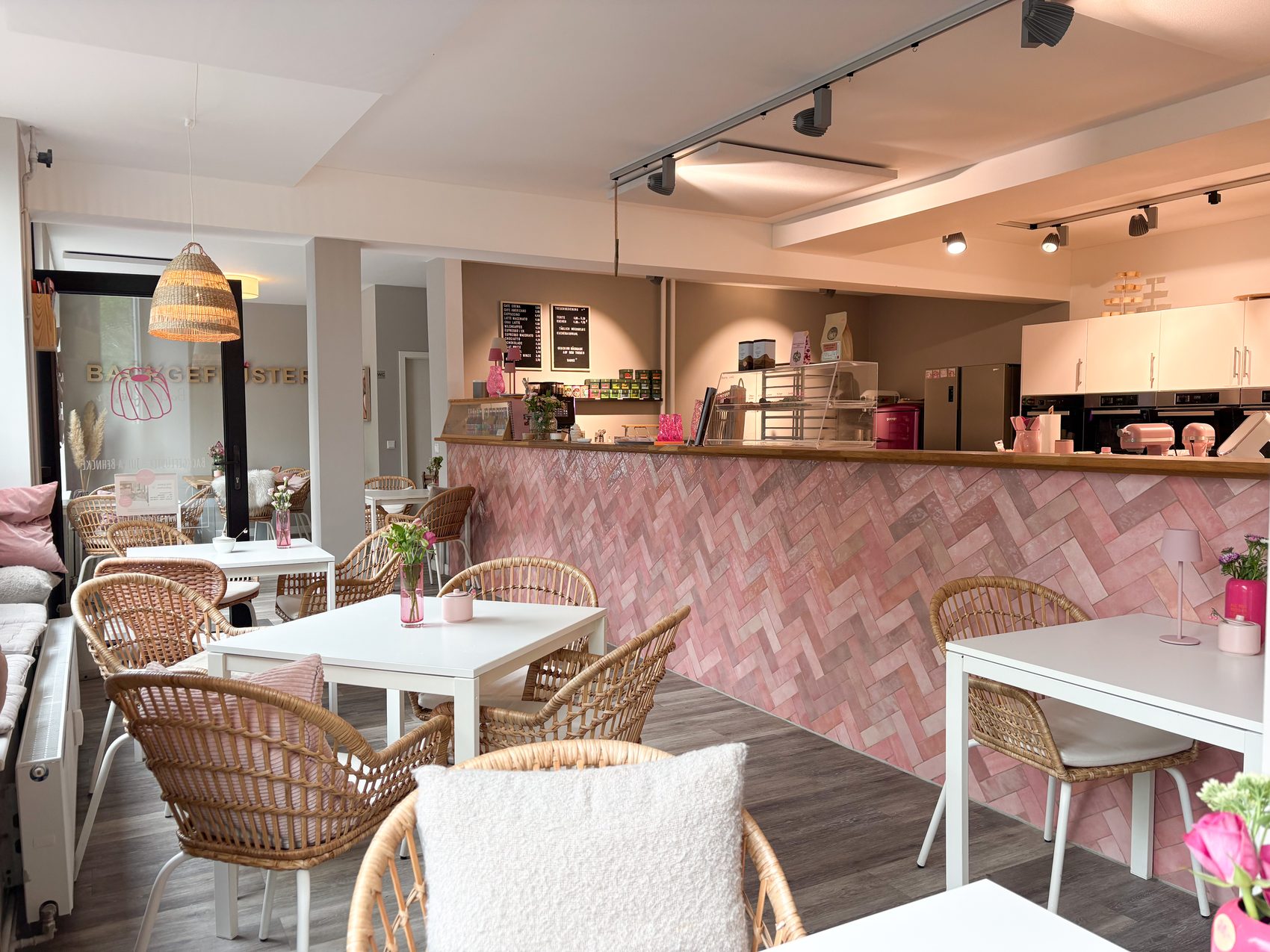 Heller Gastraum im Café Backgeflüster mit Rattanstühlen, weißen Tischen und rosa Tresen im Hintergrund