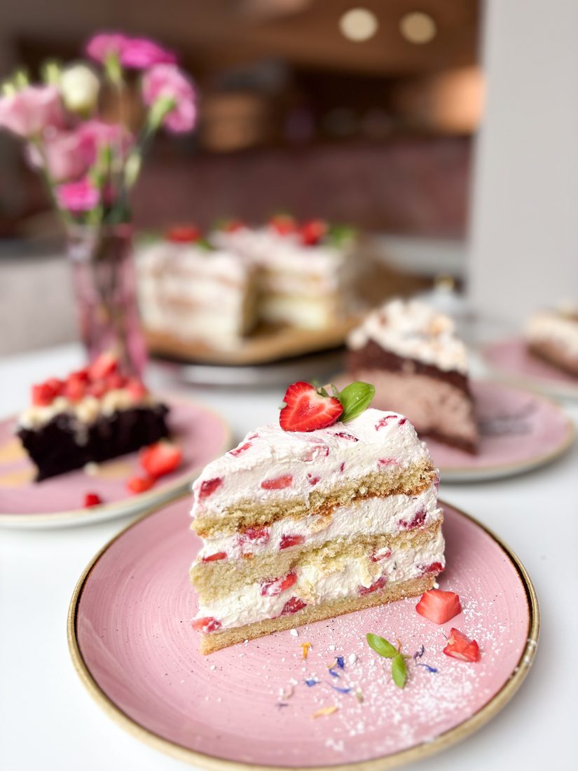 Erdbeer-Sahne-Torte auf einem rosa Teller, dahinter weitere Kuchen